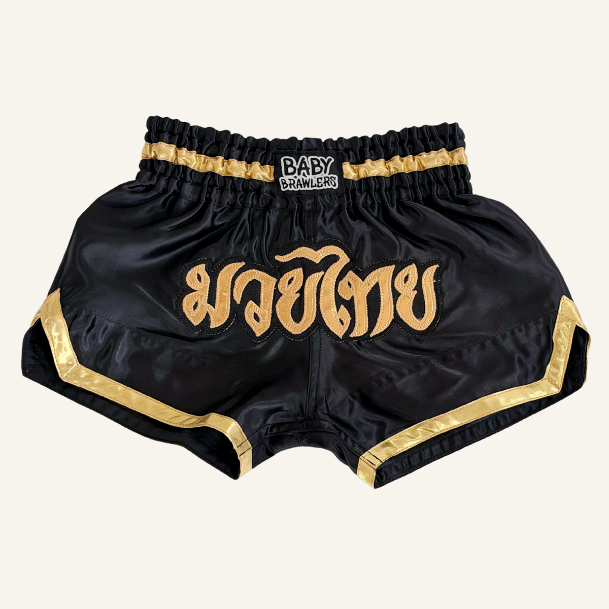 Black satin Muay Thai shorts with gold trim and Thai script embroidery on the front, featuring a black elastic waistband with a 'BABY BRAWLERS' logo patch.
