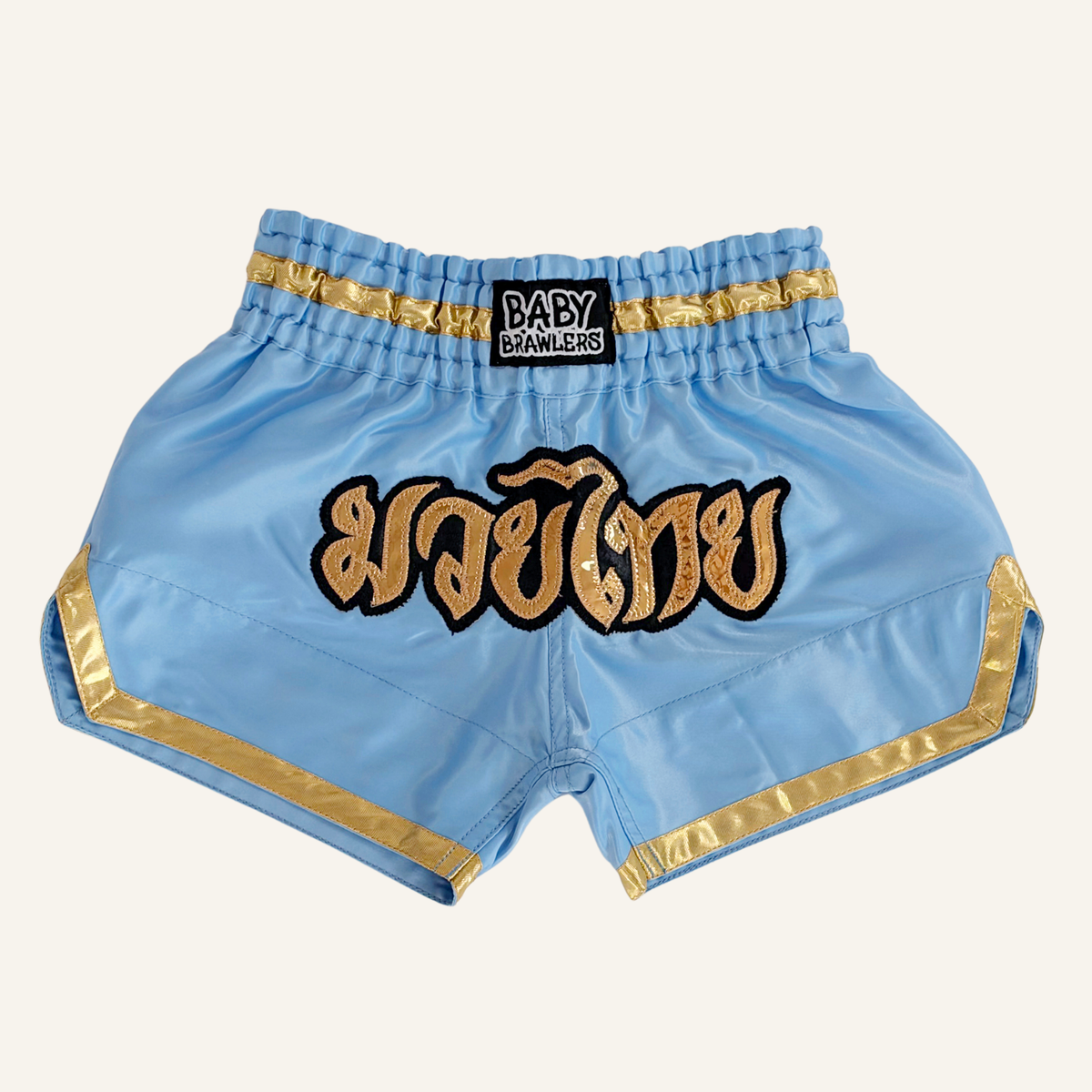 Light blue satin Muay Thai shorts with gold trim, featuring a black waistband patch with 'BABY BRAWLERS' text and a large gold embroidered Thai script patch on the front.