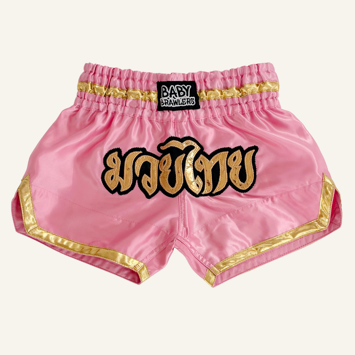 Pink satin Muay Thai shorts with gold and black trim, featuring a large gold Thai script patch on the front and a black waistband patch reading 'BABY BRAWLERS'.
