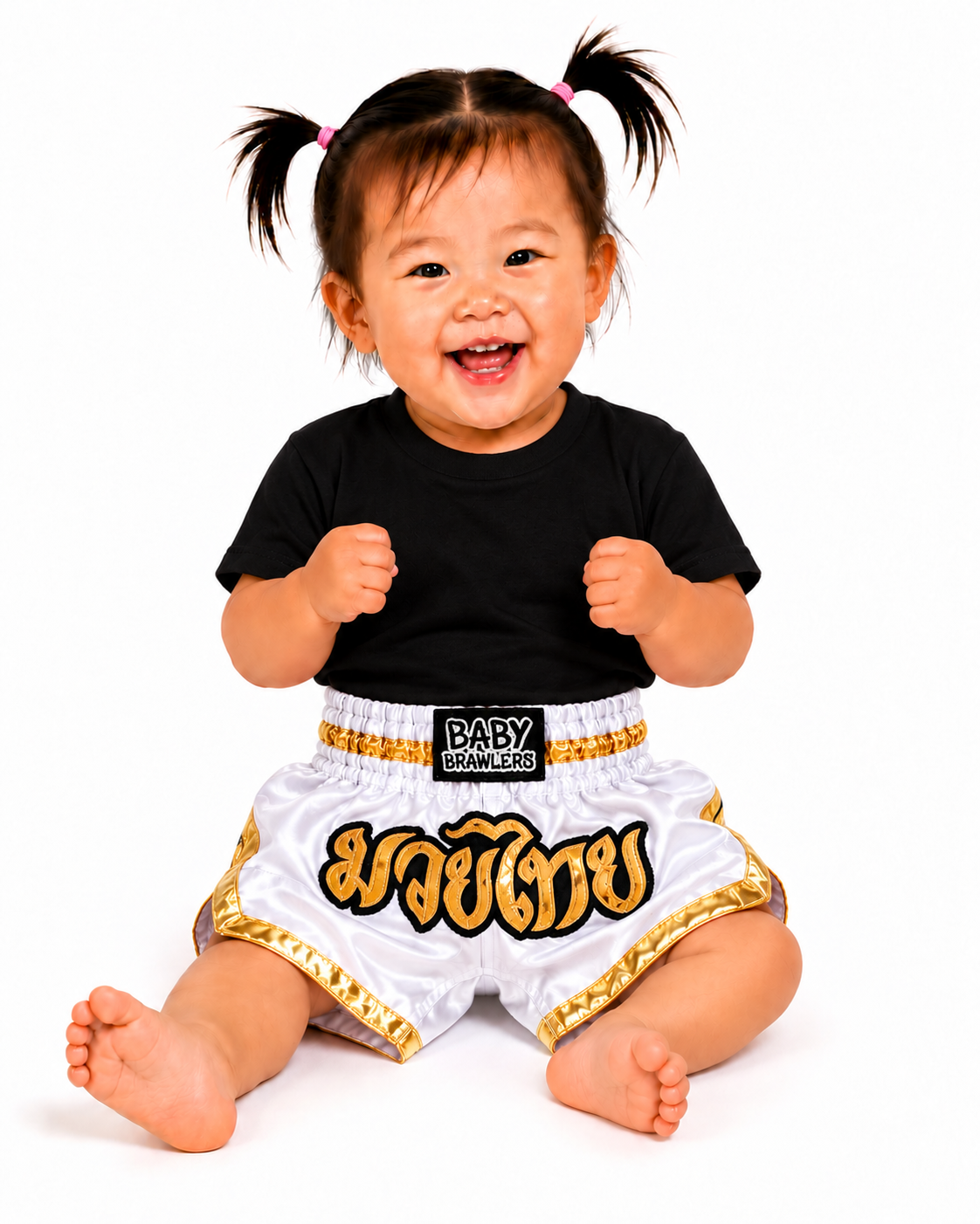 A smiling baby sitting on a white background wearing white Muay Thai shorts with gold trim and a black waistband patch that reads 'BABY BRAWLERS'.